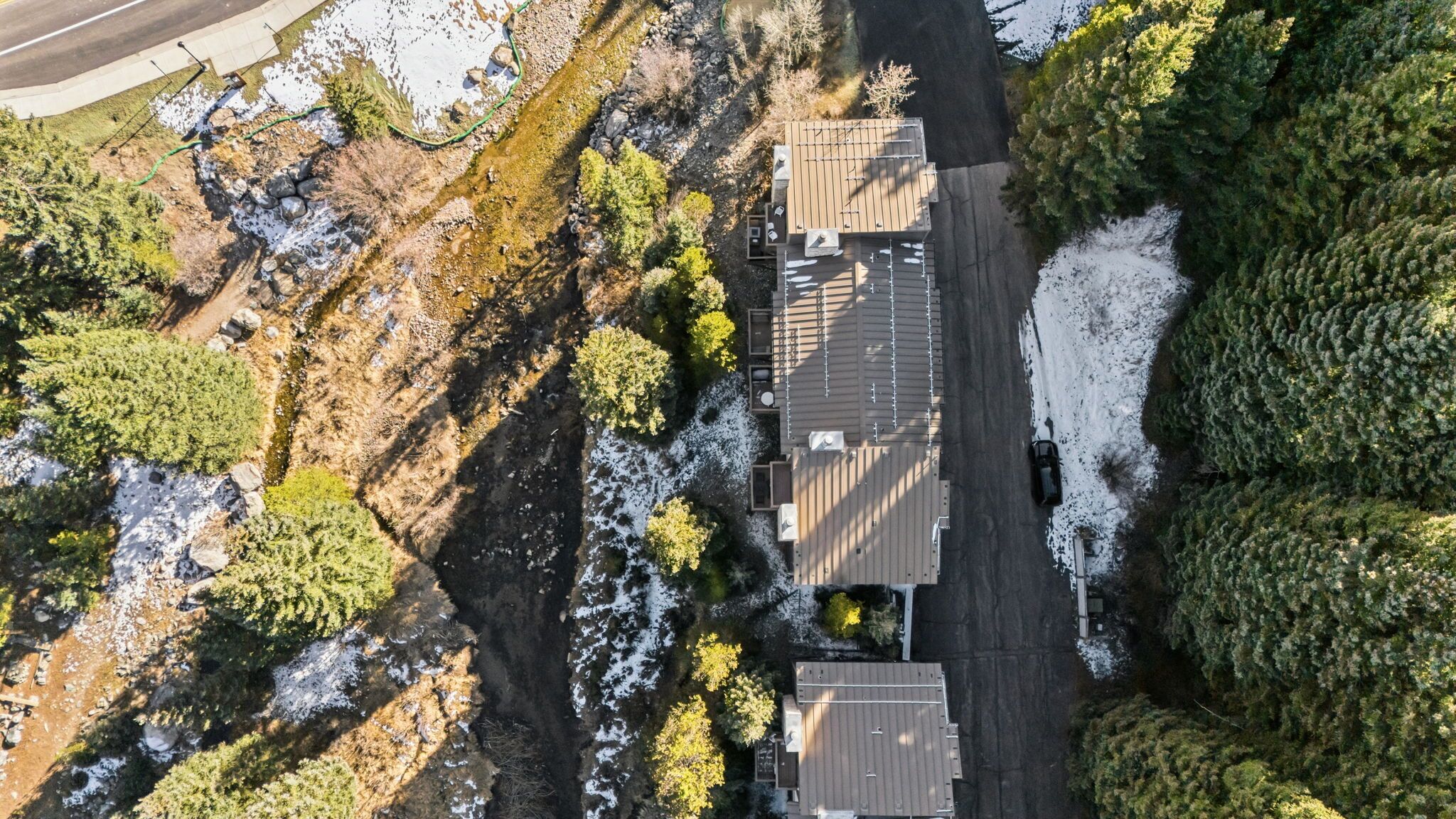 An aerial view of a residential area with a house featuring a sloped roof, a driveway, and a small yard with trees and a snow-covered ground.
