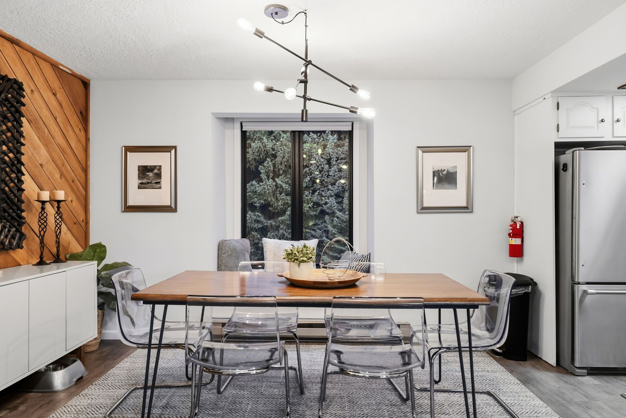 Une cuisine moderne avec une table à manger en bois, des chaises transparentes, un luminaire suspendu et un réfrigérateur.