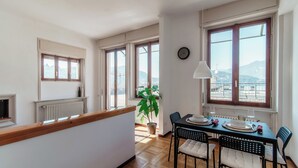A dining area with a table set for two, large windows with a view of mountains, a potted plant, and a radiator. - Terrazza Rooftop (Como)