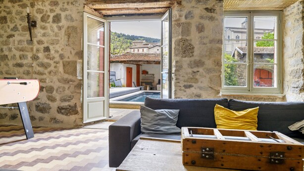 Una habitación con pared de piedra, una mesa de madera, un sofá, una maleta y vistas a una piscina y edificios.