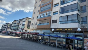 A modern building with multiple floors, a sign for 'House Beach', and outdoor seating with umbrellas.