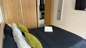 A neatly made bed with a dark bedspread and pillows, a wooden wardrobe, and a flat-screen TV mounted on the wall.