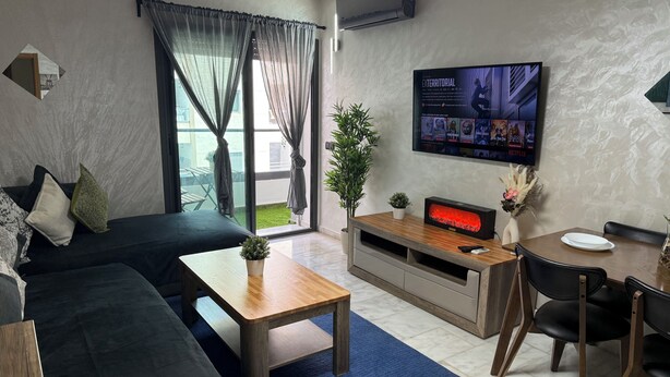 A modern living room with a dark-colored sofa, a wooden coffee table, a TV on a wooden stand, and a dining area with a table and chairs.