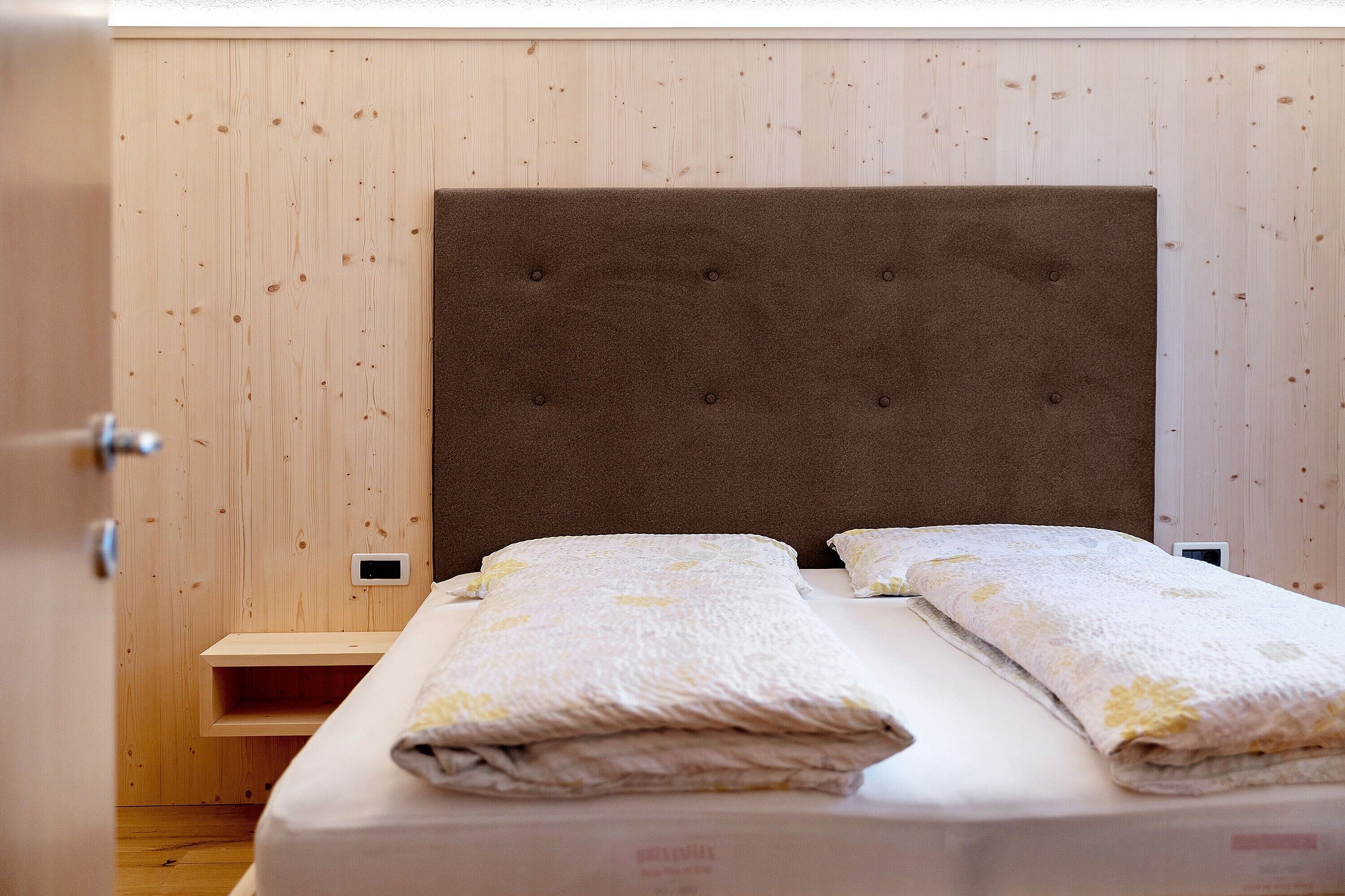 A neatly made bed with a headboard, a bedside table, and a wall-mounted thermostat.