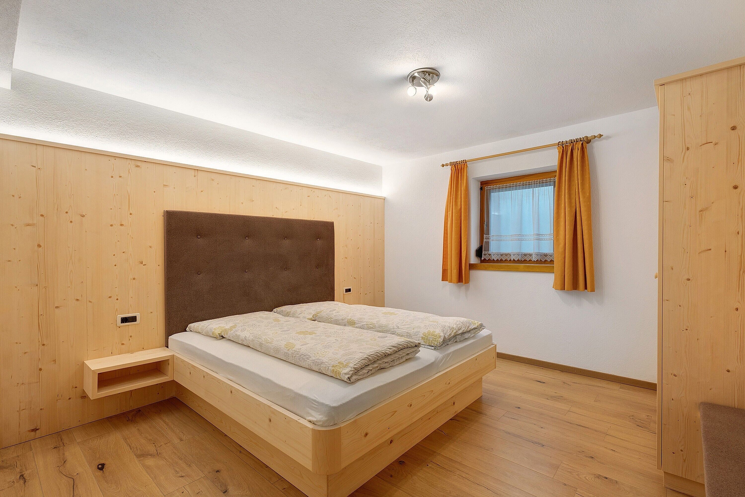 A bedroom with a wooden bed, a built-in shelf, a window with curtains, and a wardrobe.