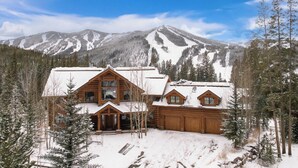 A log cabin with a large porch and multiple windows, surrounded by snow-covered trees and mountains. - Forest Setting | Near Trail Access (Winter Park)