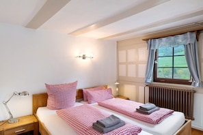 A double bed with checkered bedding, a bedside table with a lamp, a window with curtains, and a radiator.