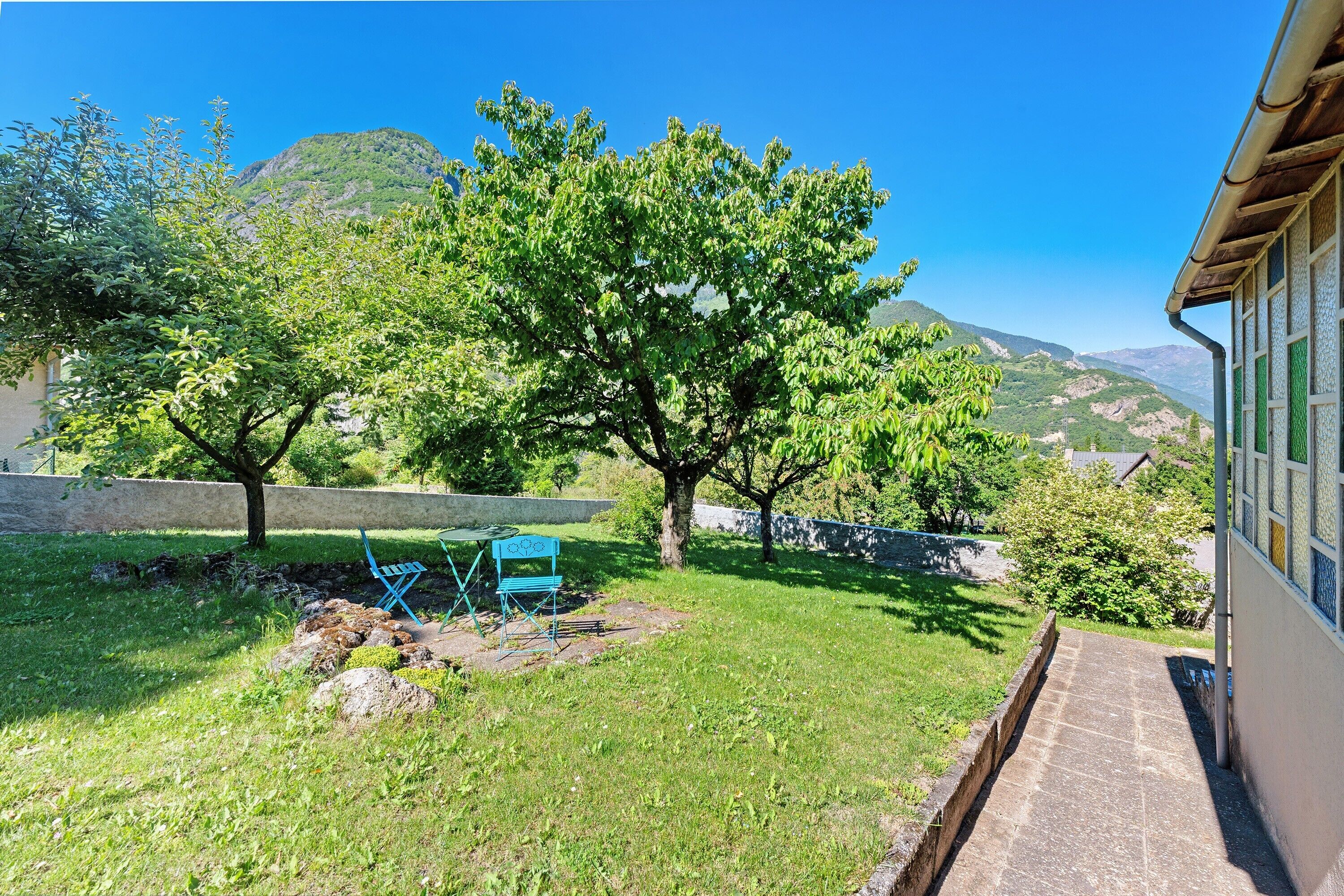 A grassy backyard with a tree, a blue chair, and a stone wall.