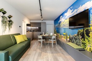 Een moderne leefruimte met een groene bank, een eettafel met stoelen, een keukenhoek en een grote televisie aan een muur met een sfeervol muurschildering.