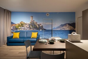 Una stanza con un ampio murale panoramico, un divano blu con cuscini gialli, un tavolo in legno con piatti e ciotole, e un 'Welcome Kit' sul tavolo.