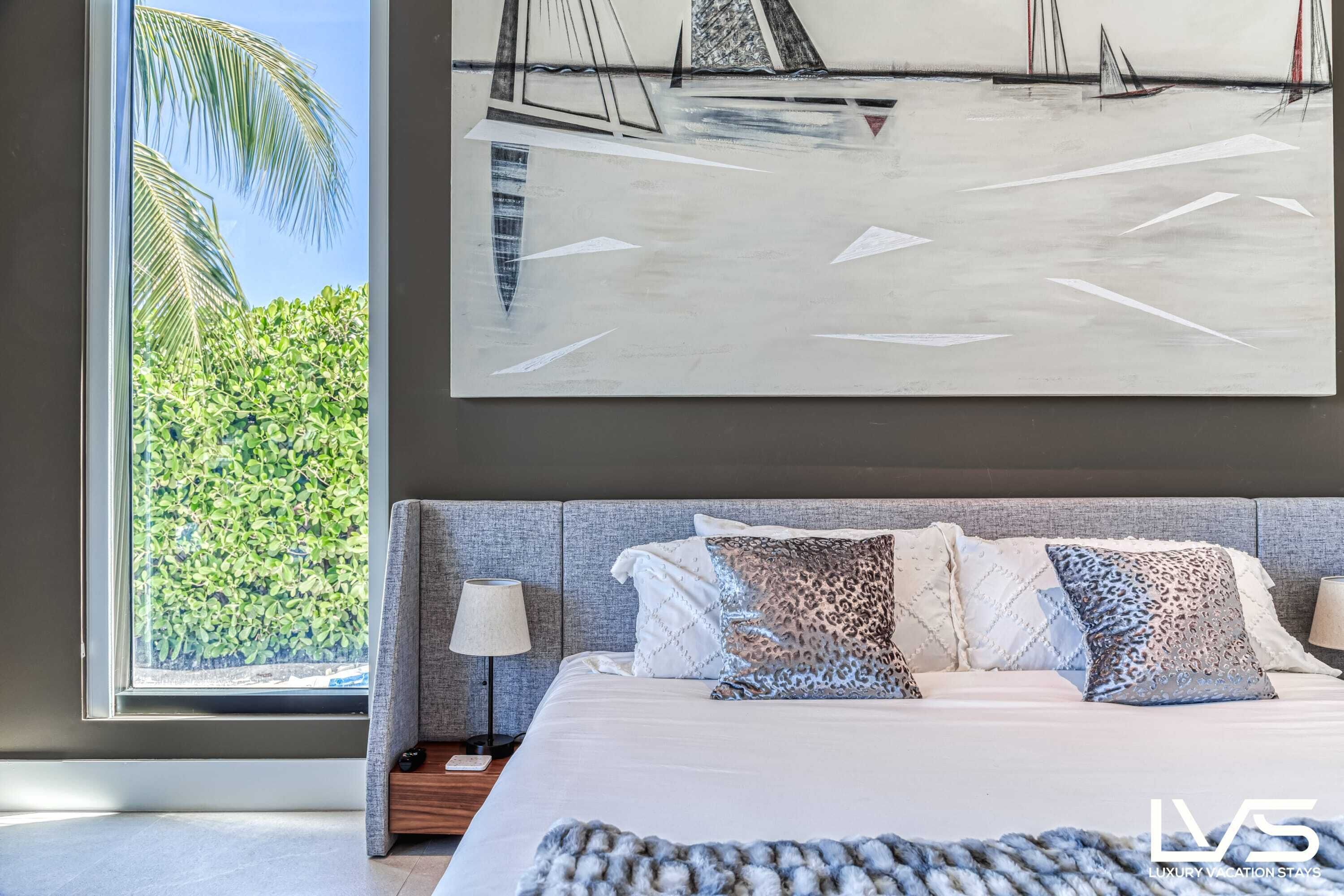 A bedroom with a large bed, a view of greenery through a window, and a painting of sailboats on the wall.