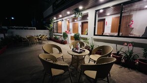 An outdoor seating area with wicker chairs and tables, surrounded by potted plants and a building with large windows. - Hotel Gulmarg Residency (Mount Abu)