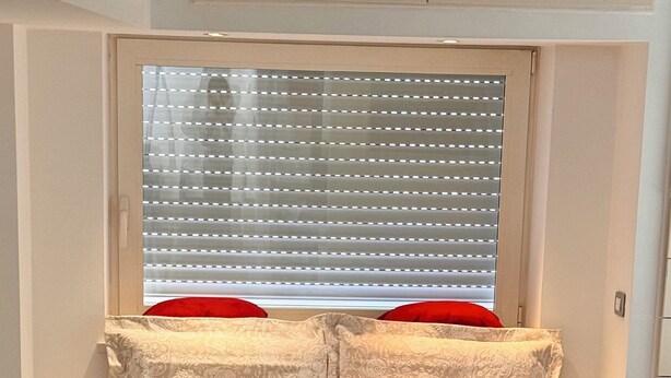 En snyggt bäddad säng med vita sängkläder och en mönstrad sänggavel, ett fönster med persienner och ett klinkergolv.