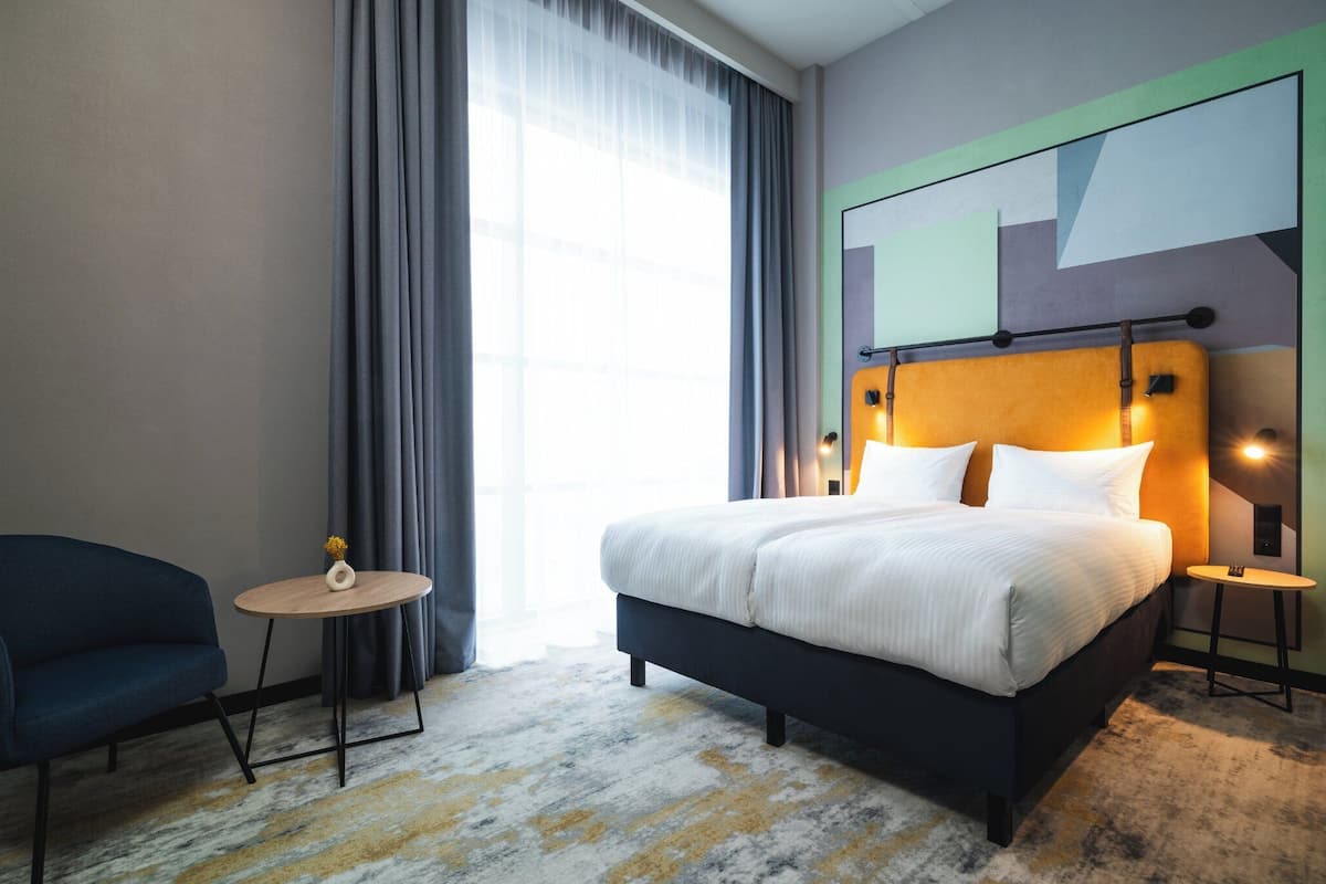 Habitación de hotel moderna con una cama grande, una mesita redonda pequeña, una silla y vistas de la ciudad a través del ventanal.