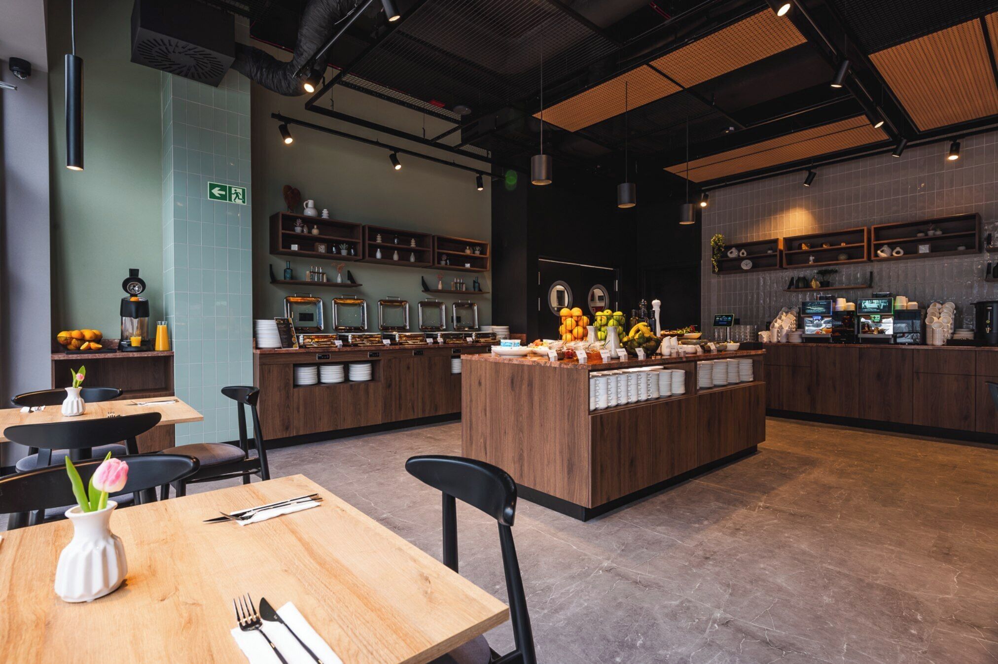 L’intérieur d’un restaurant présente des tables et des chaises en bois, un comptoir central où sont exposés des produits alimentaires, et des étagères remplies d’ustensiles de cuisine.