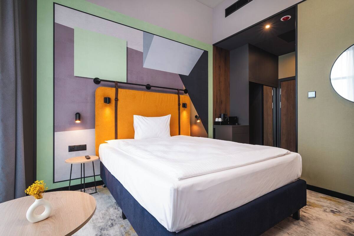 Una habitación de hotel moderna con una cama grande, una mesita redonda con un florero y un diseño geométrico único en la pared.