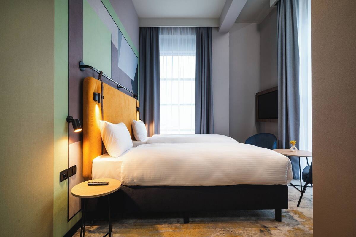 Habitación de hotel con una cama grande, una mesita de noche con control remoto, una pequeña mesa redonda y un televisor sobre un soporte.