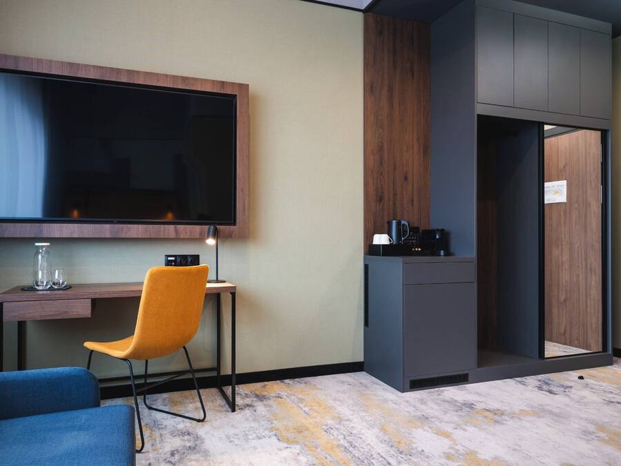 Ett modernt hotellrum med en stor platt-TV som är monterad på väggen, ett skrivbord med en stol, en kaffemaskin och en garderob med en spegel.
