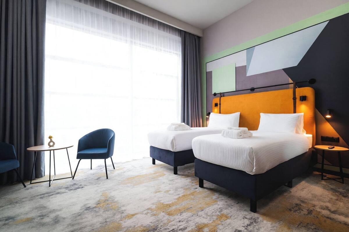 Una habitación de hotel con dos camas, una mesita, dos sillas y un ventanal con cortinas.