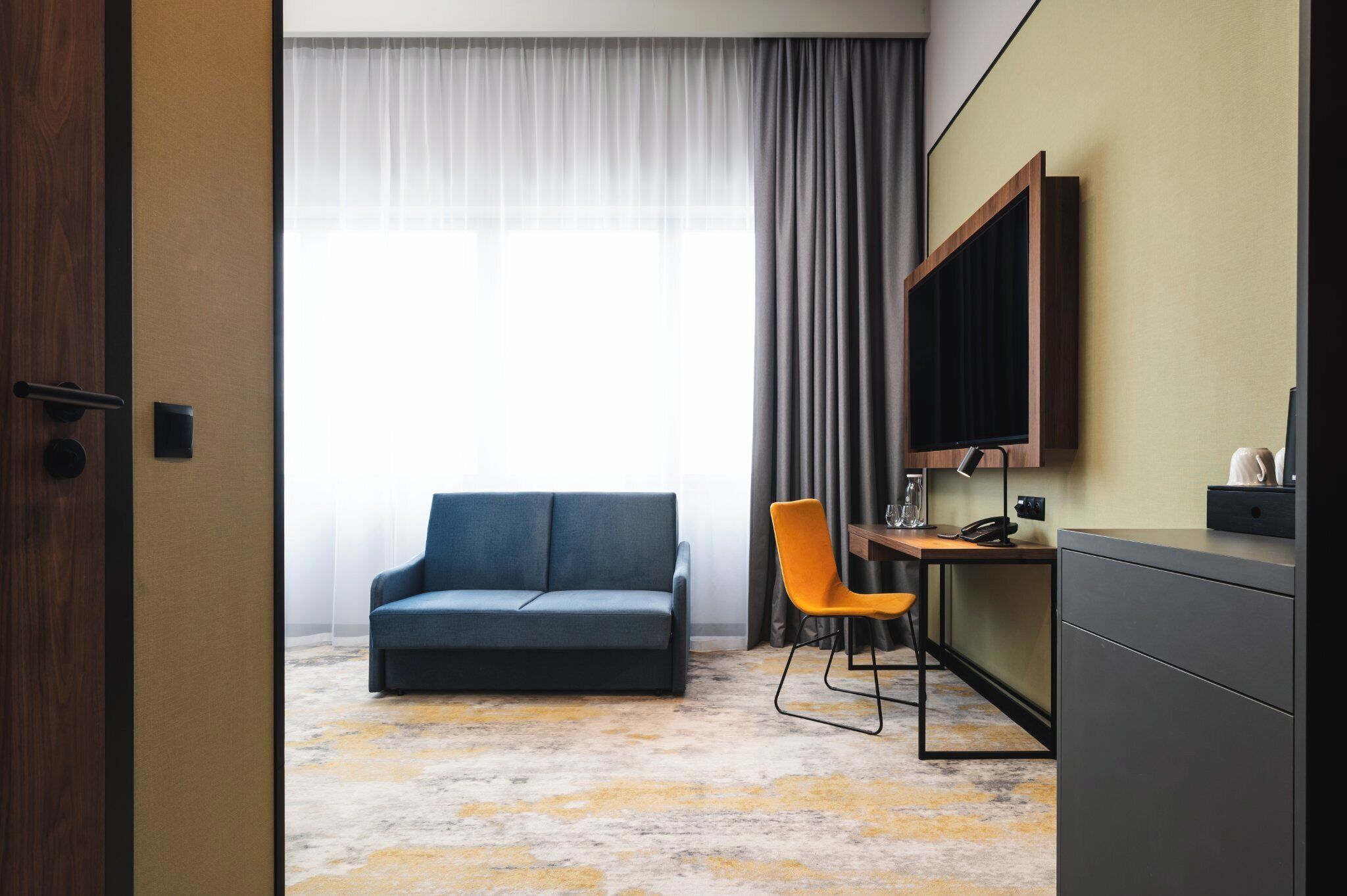 A hotel room with a sofa, a desk, a chair, a TV, and a large window with curtains.