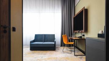 A hotel room with a sofa, a desk, a chair, a TV, and a large window with curtains.