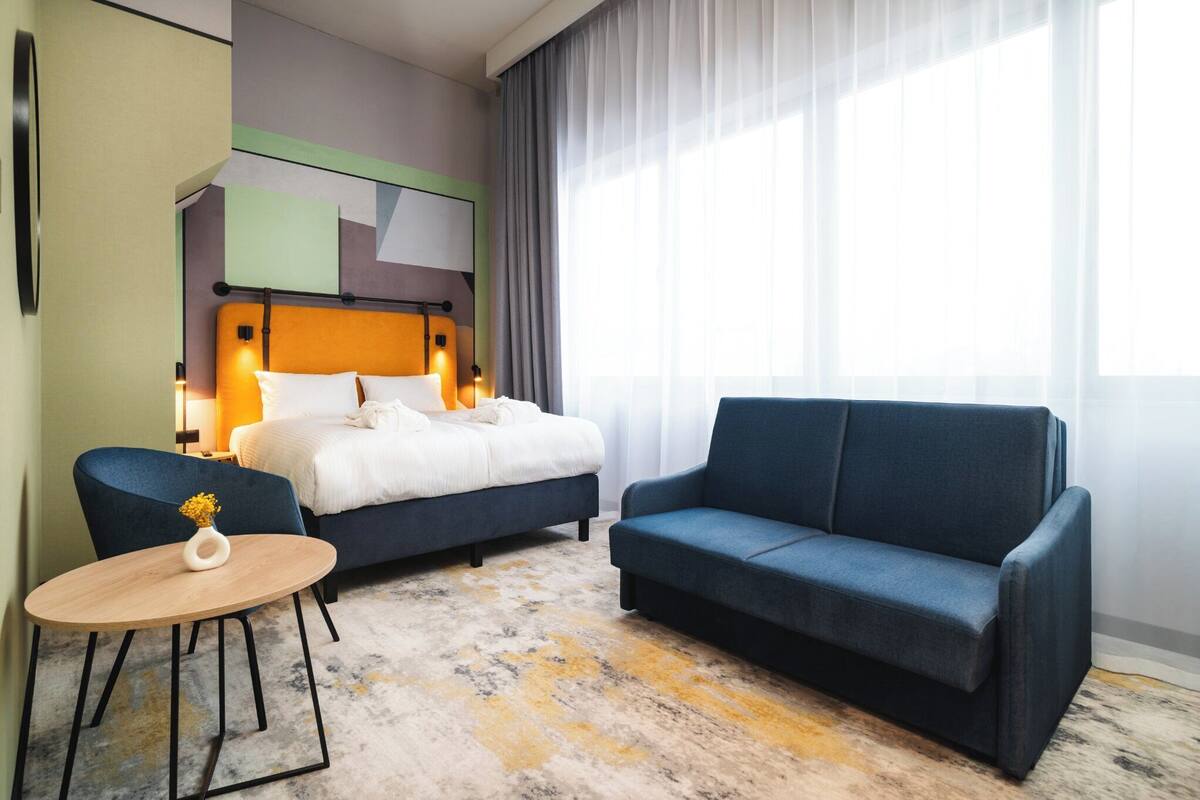 Una habitación de hotel con una cama, un sofá azul, una mesita con un jarrón y un ventanal con cortinas.