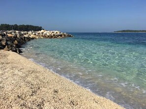Una playa de guijarros con agua cristalina, un rompeolas rocoso y una masa de tierra cubierta de árboles en la distancia.