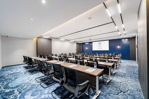 Un salón de conferencias con filas de mesas y sillas, una pantalla de proyector y una alfombra azul.