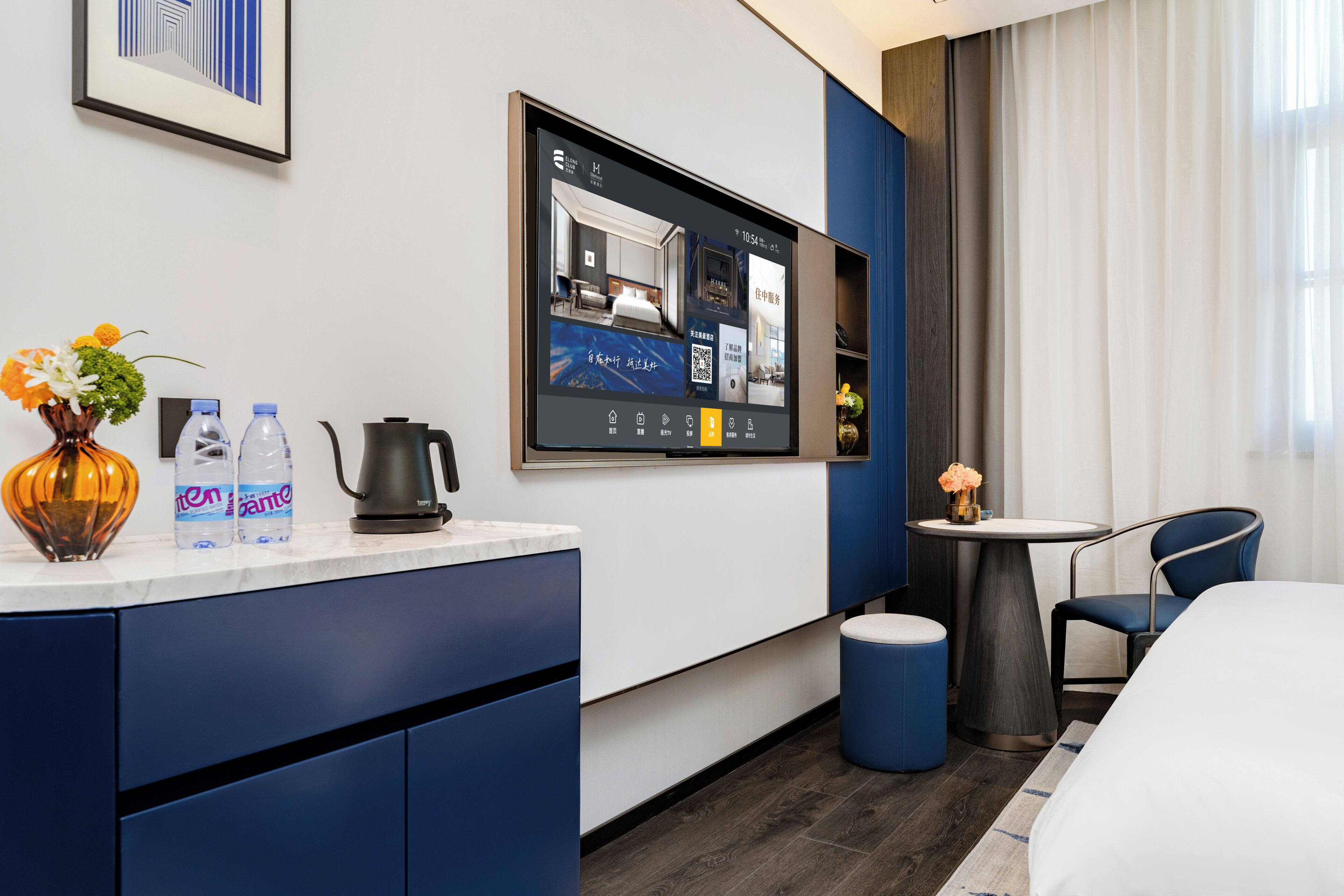 A hotel room with a bed, a desk with a computer monitor, a coffee maker, water bottles, and a vase with flowers.