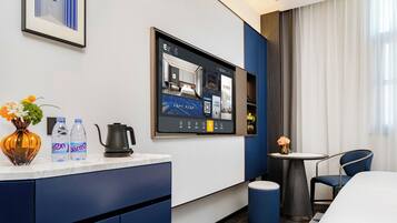 A hotel room with a bed, a desk with a computer monitor, a coffee maker, water bottles, and a vase with flowers.