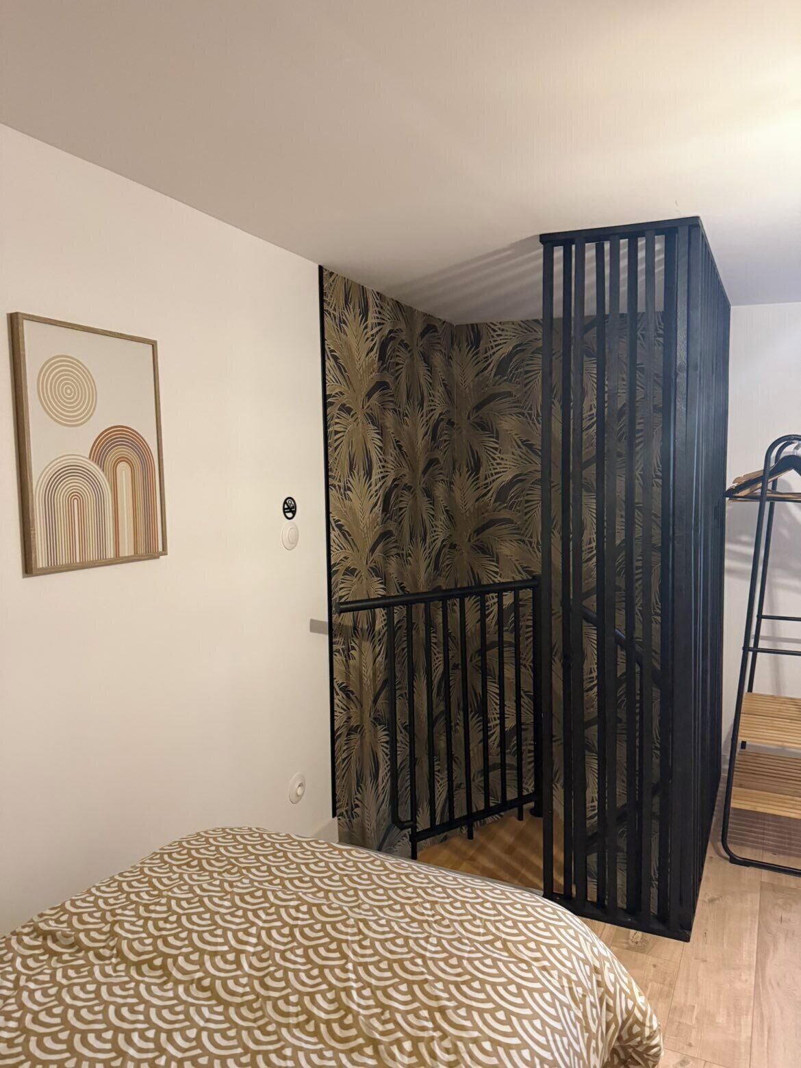 A bedroom with a bed, a wall with a circular artwork, a door with a palm leaf pattern, and a black metal railing.