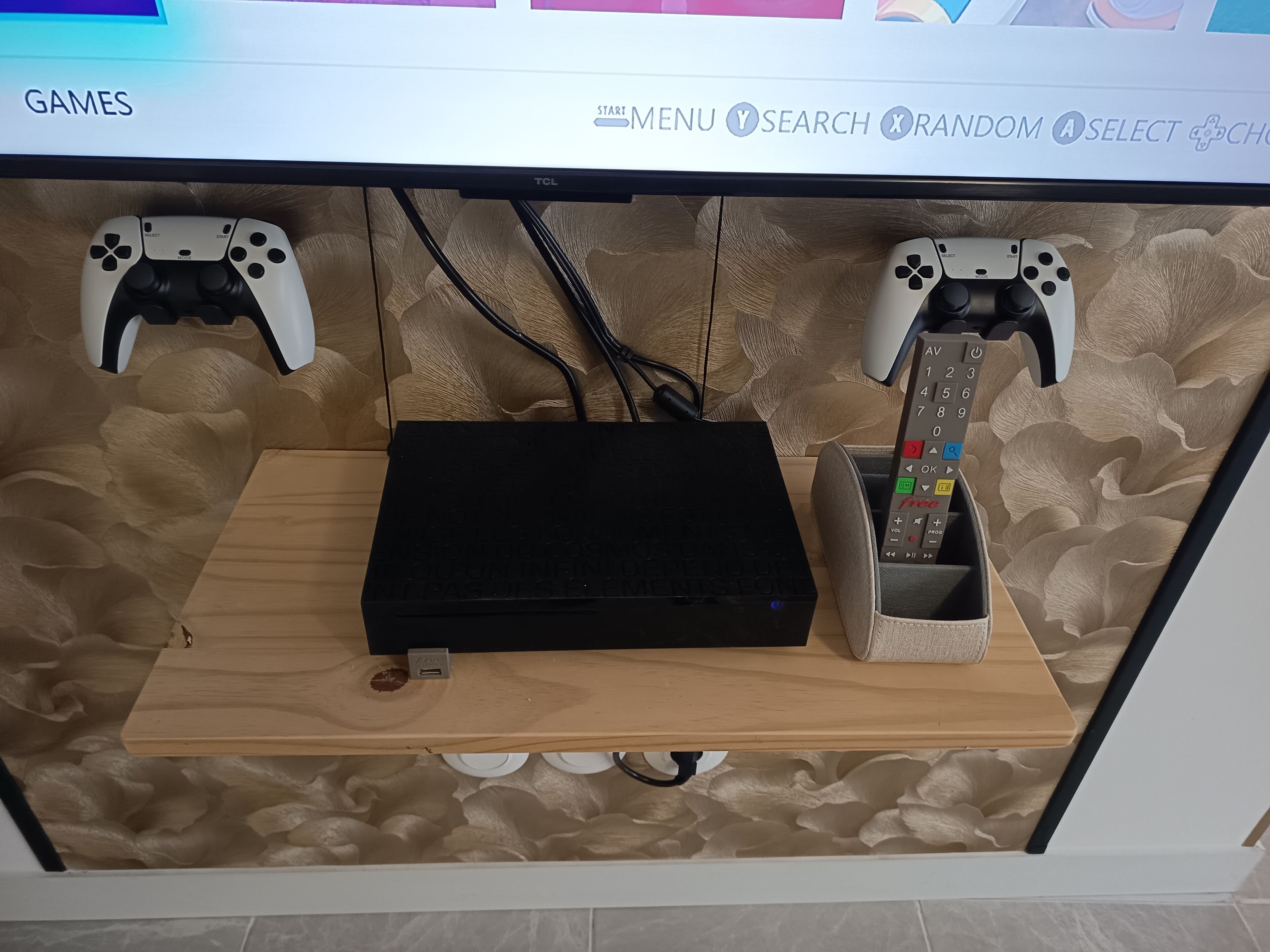 En speluppställning med två handkontroller, en router och en fjärrkontroll på en hylla i trä.