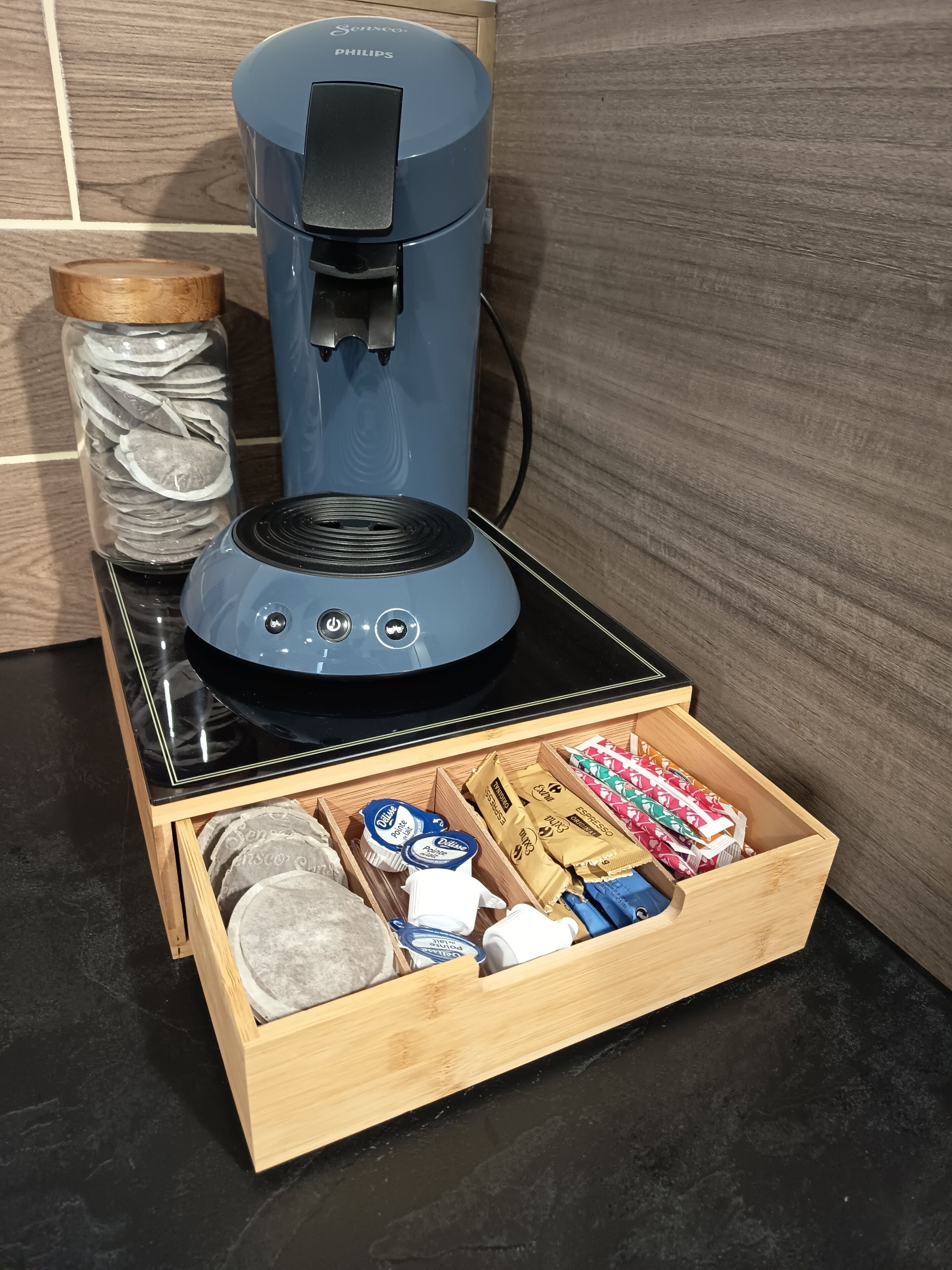 Une machine à café posée sur un plan de travail noir, avec un tiroir contenant des dosettes de café et un récipient rempli de café moulu.