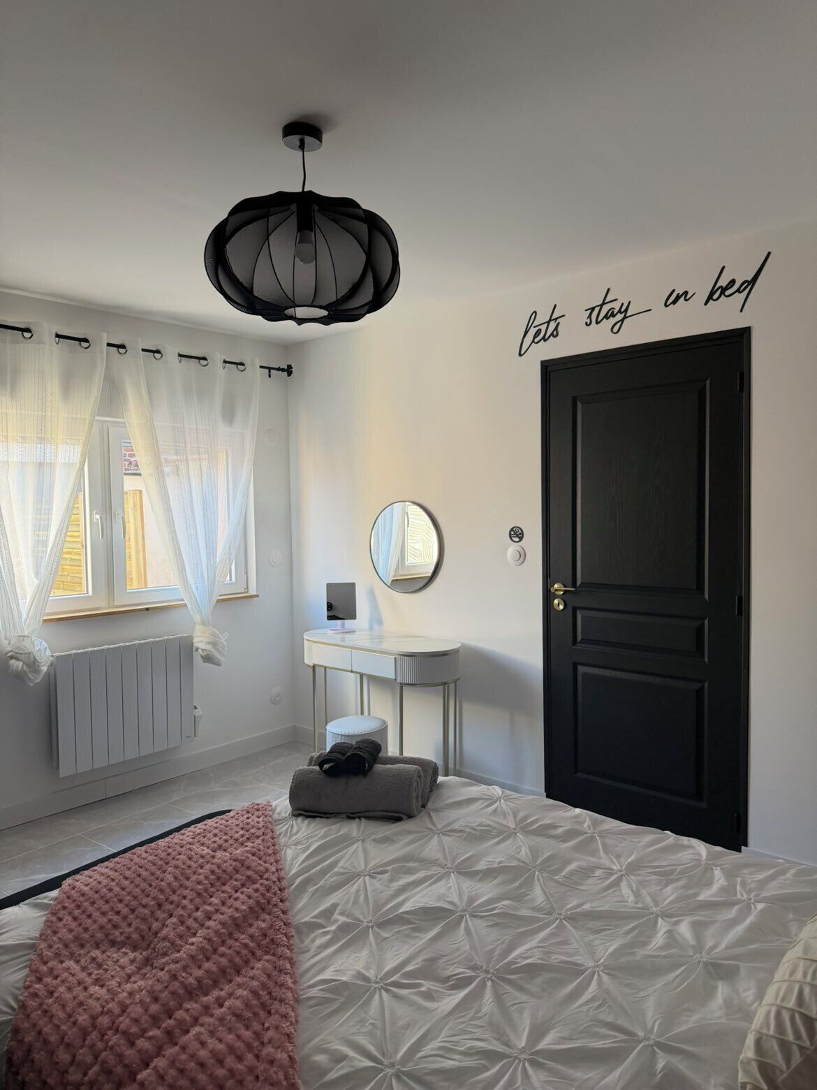 A bedroom with a bed, a bedside table, a mirror, a black door, and a window with sheer curtains.