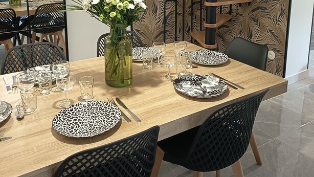 Un coin repas avec une table en bois dressée pour six personnes, entourée de chaises noires. Un vase rempli de fleurs est posé sur la table.