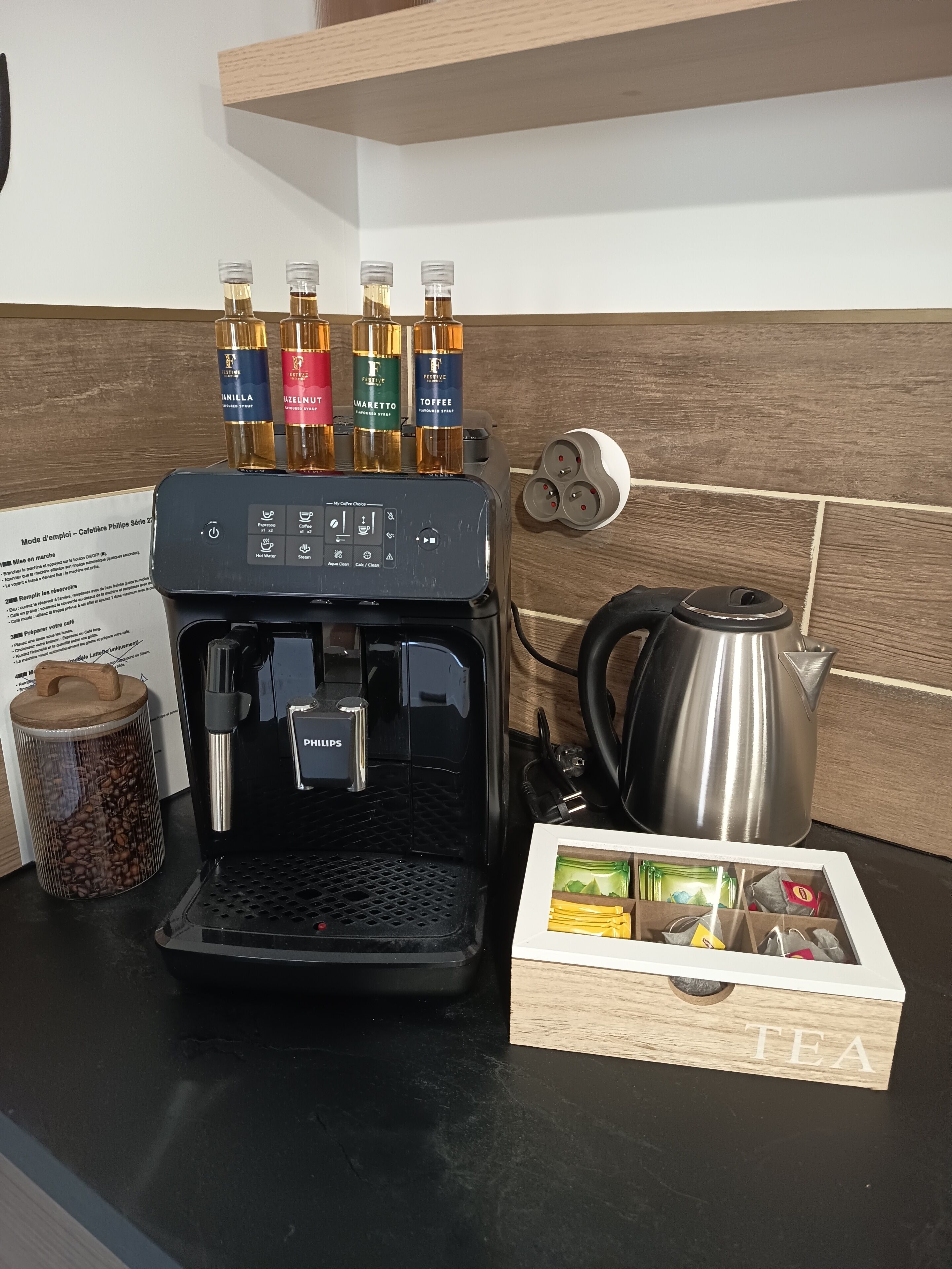 Uma máquina de café com quatro garrafas de xarope, uma chaleira, um moedor de café e uma caixa com a inscrição 'TEA'.
