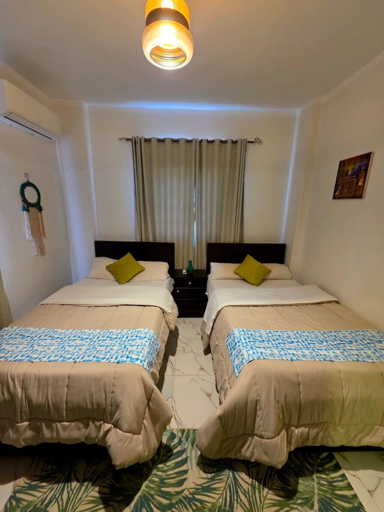 Une chambre d’hôtel avec deux lits, un luminaire au plafond, une fenêtre avec des rideaux, un climatiseur fixé au mur et une petite table sur laquelle se trouve une bouteille verte.