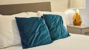 Ein Bett mit weißen Leinen und türkisfarbenen Kissen, ein Nachttisch mit Lampe und ein graues Kopfstück.