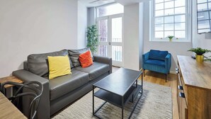 Ein modernes Wohnzimmer mit einem grauen Sofa, einem Couchtisch, einem blauen Sessel und einem Holzesstisch. Große Fenster sorgen für natürliches Licht und bieten einen Blick auf die Stadt.