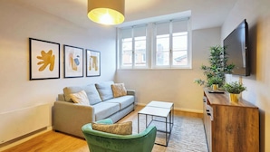 Un salon avec un canapé, un fauteuil vert, une table basse, un téléviseur et des œuvres d’art encadrées accrochées au mur.