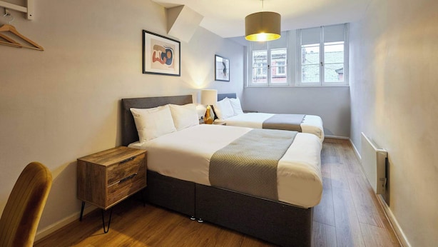 Une chambre avec deux lits, une commode en bois, un bureau et une chaise. Deux fenêtres laissent entrer la lumière naturelle.