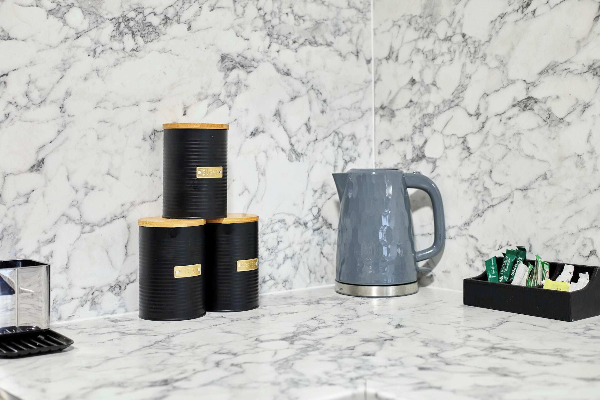 A marble countertop with black containers stacked on top of each other, a grey pitcher, and a black tray with green items.