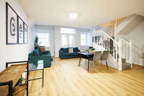 Amplia sala de estar con piso de madera, una escalera con barandillas blancas y un comedor con mesa y sillas.