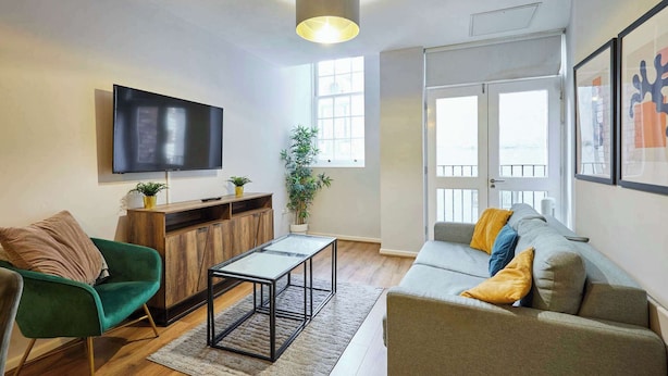 Een moderne woonkamer met een flatscreen televisie, een houten kast, een groene stoel, een grijze bank met gele kussens en een groot raam.