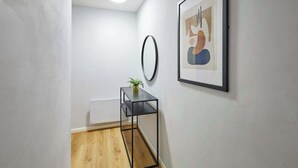 Un passadís amb sòl de fusta, una taula auxiliar de metall negre amb una petita planta, un mirall rodó i un quadre abstracte amb marc a la paret.