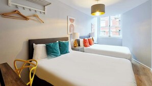 Habitación de hotel con dos camas, un perchero, mesitas de noche con lámparas y una ventana con vista a edificios.