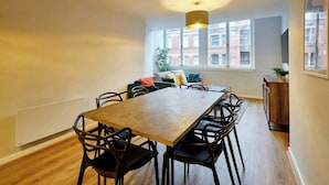 Un comedor con una mesa y sillas de madera, una lámpara colgante, un ventanal con vistas a edificios y una planta en el alféizar.