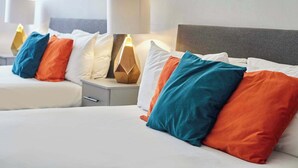 Ein Hotelzimmer mit zwei Betten, jeweils mit weißer Bettwäsche und mehreren Kissen in Blau, Orange und Weiß. Es gibt Nachttischlampen und einen Nachttisch mit einer Lampe.