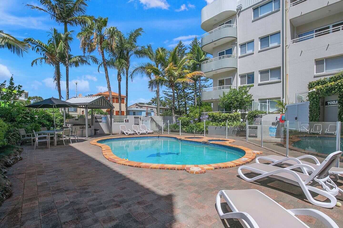 A swimming pool with lounge chairs, surrounded by palm trees and a building with balconies.