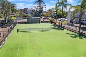 En tennisbane med net, omgivet af et hegn, beliggende ved et boligområde med huse og parkerede biler.